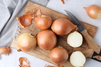 Fresh onions with peels and knife on white tiled table, top view