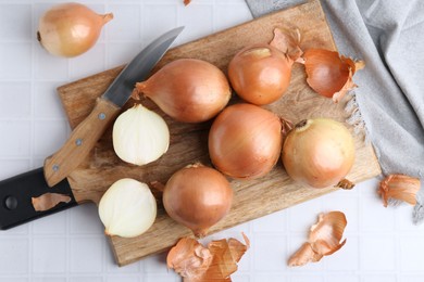 Fresh onions with peels and knife on white tiled table, top view