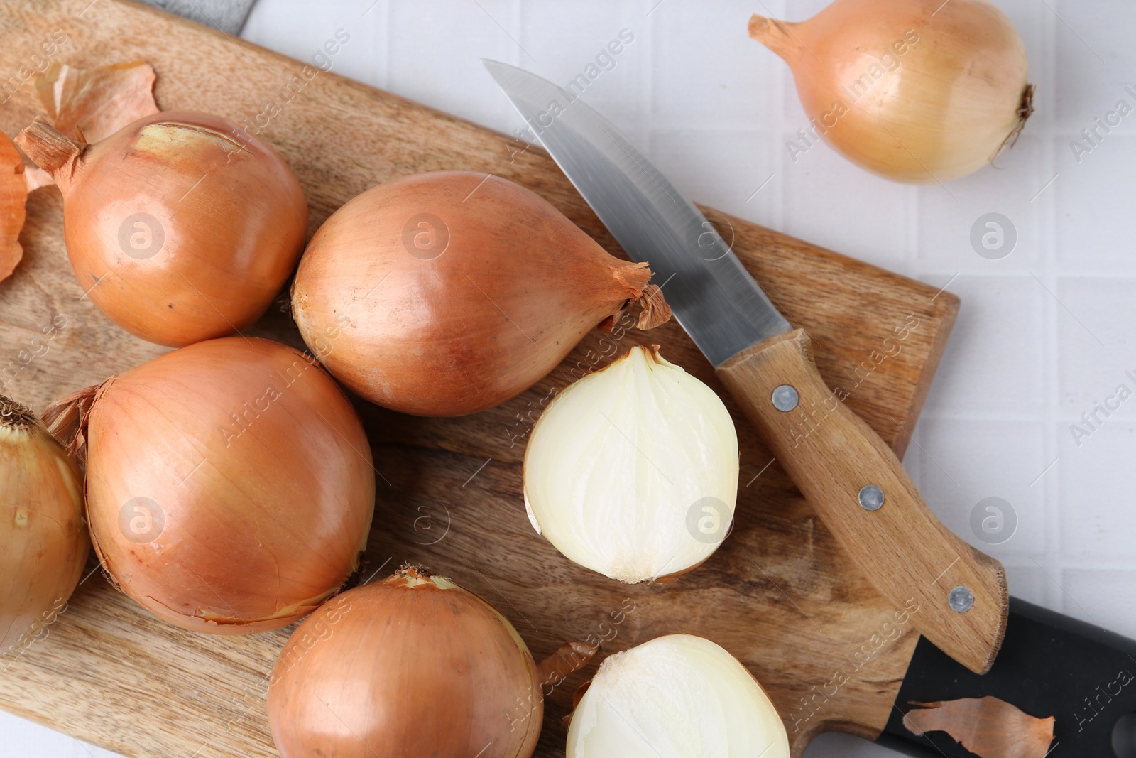 Photo of Fresh onions with peels and knife on white tiled table, top view