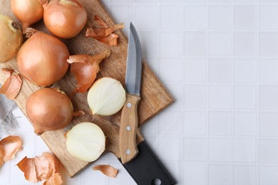 Photo of Fresh onions with peels and knife on white tiled table, top view. Space for text