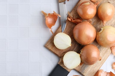 Fresh onions with peels and knife on white tiled table, top view. Space for text