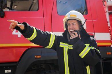 Photo of Firefighter in uniform using portable radio set near fire truck outdoors