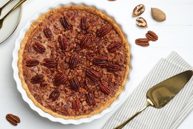 Photo of Delicious pecan pie in baking dish, fresh nuts and cake server on white wooden table, flat lay