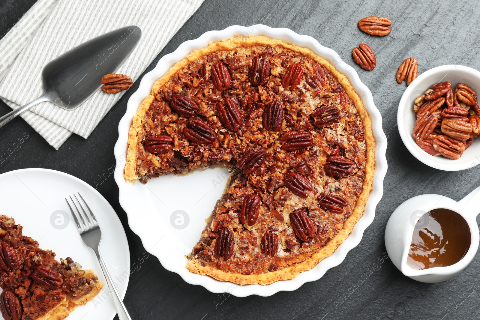 Photo of Delicious pecan pie, fresh nuts, caramel sauce and cake server on dark gray textured table, top view