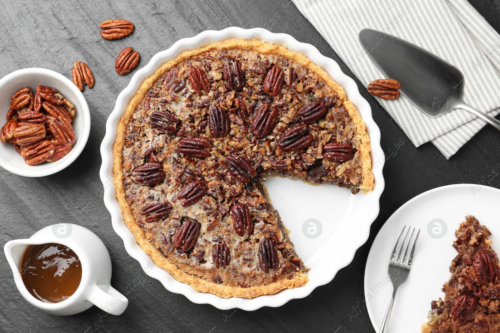 Photo of Delicious pecan pie, fresh nuts, caramel sauce and cake server on dark gray textured table, top view