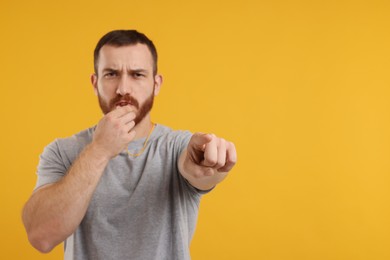 Young man blowing whistle on orange background, space for text