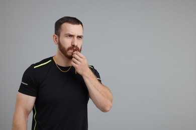 Young man blowing whistle on grey background, space for text