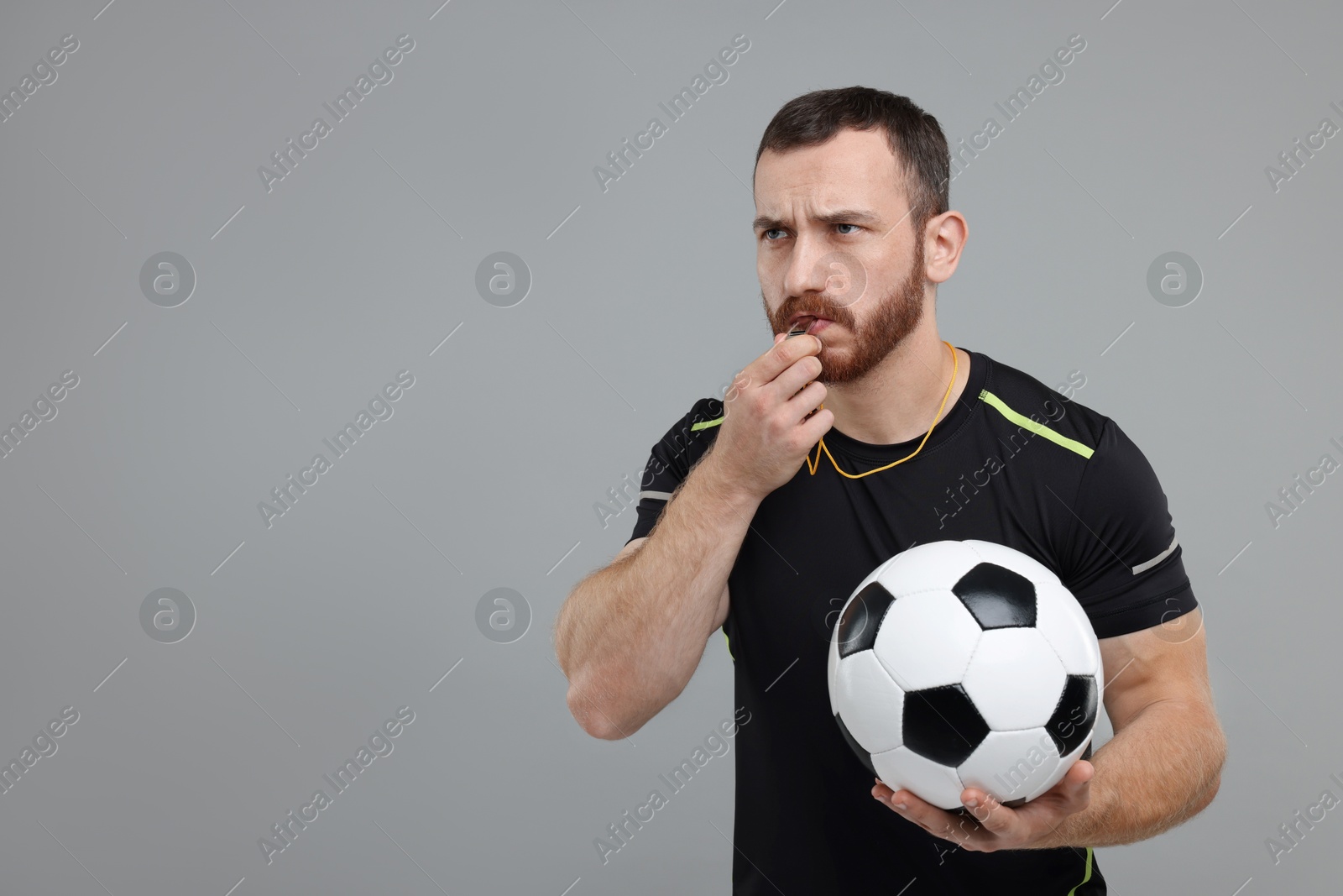 Photo of Young man with soccer ball blowing whistle on grey background, space for text