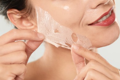 Smiling woman peeling off face mask on light grey background, closeup