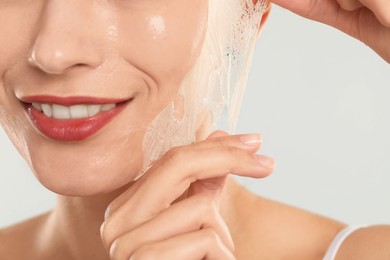 Smiling woman peeling off face mask on light grey background, closeup