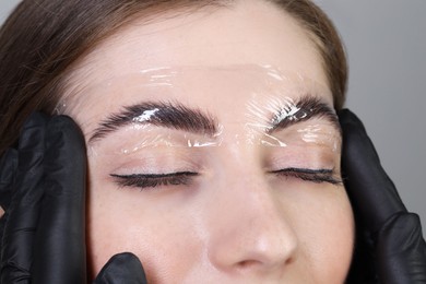 Brow lamination. Cosmetologist putting film onto woman's eyebrows against grey background, closeup