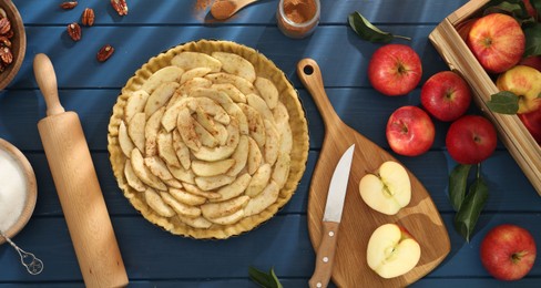 Photo of Uncooked homemade apple pie and ingredients on blue wooden table, flat lay