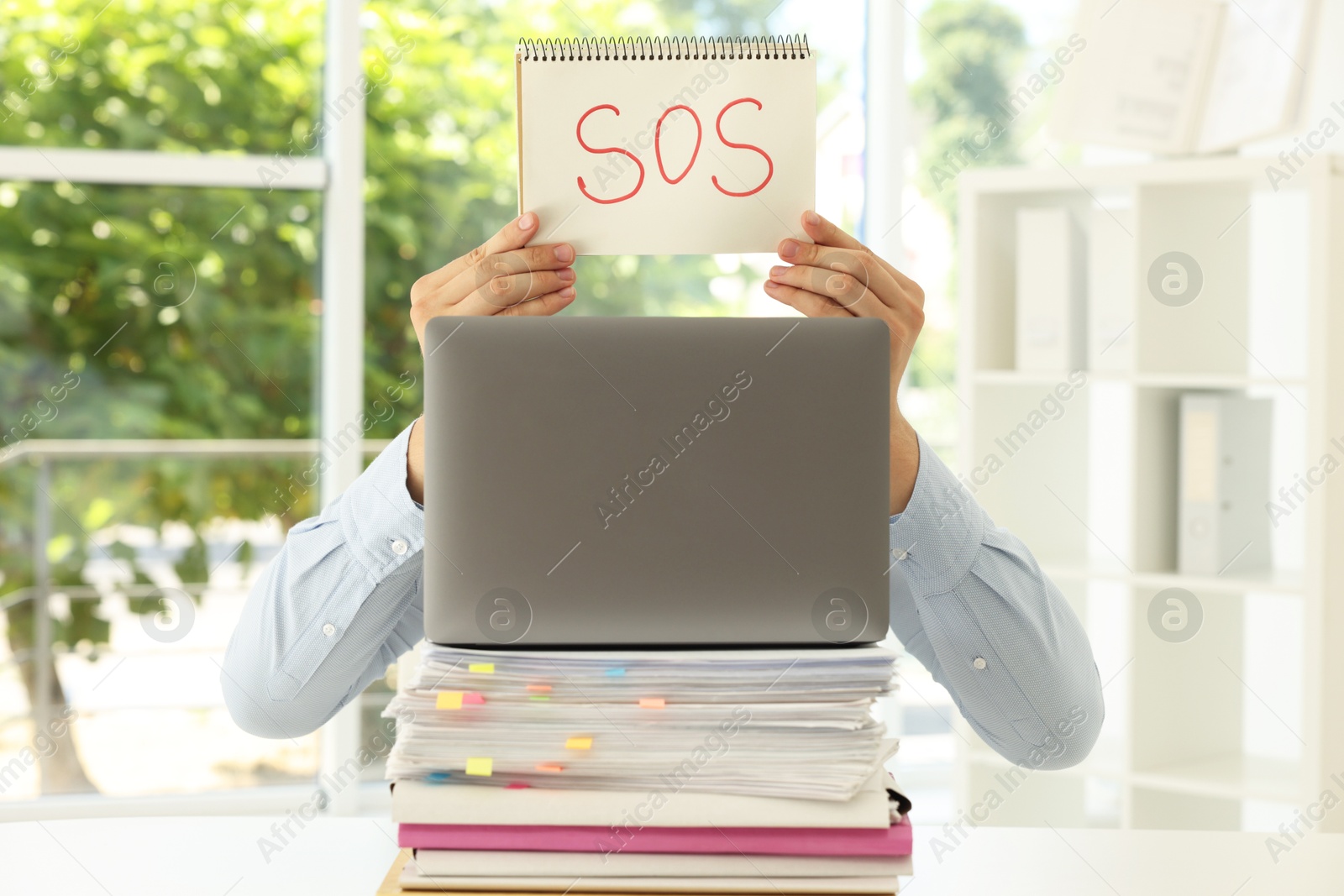 Photo of Man holding notebook with word SOS and hiding face behind laptop at table in office