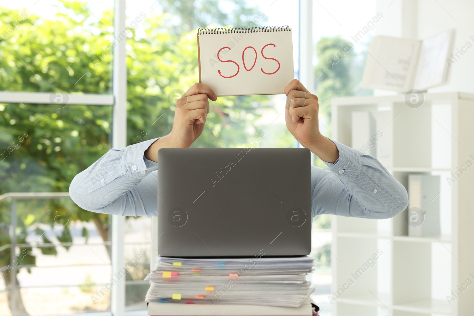 Photo of Man holding notebook with word SOS and hiding face behind laptop in office