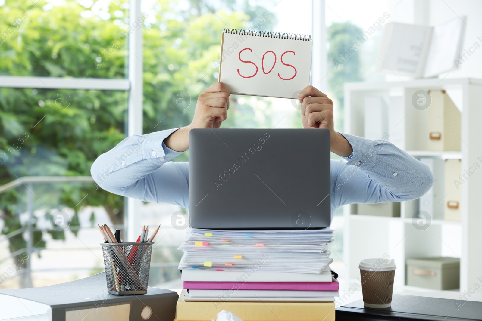 Photo of Man holding notebook with word SOS and hiding face behind laptop in office