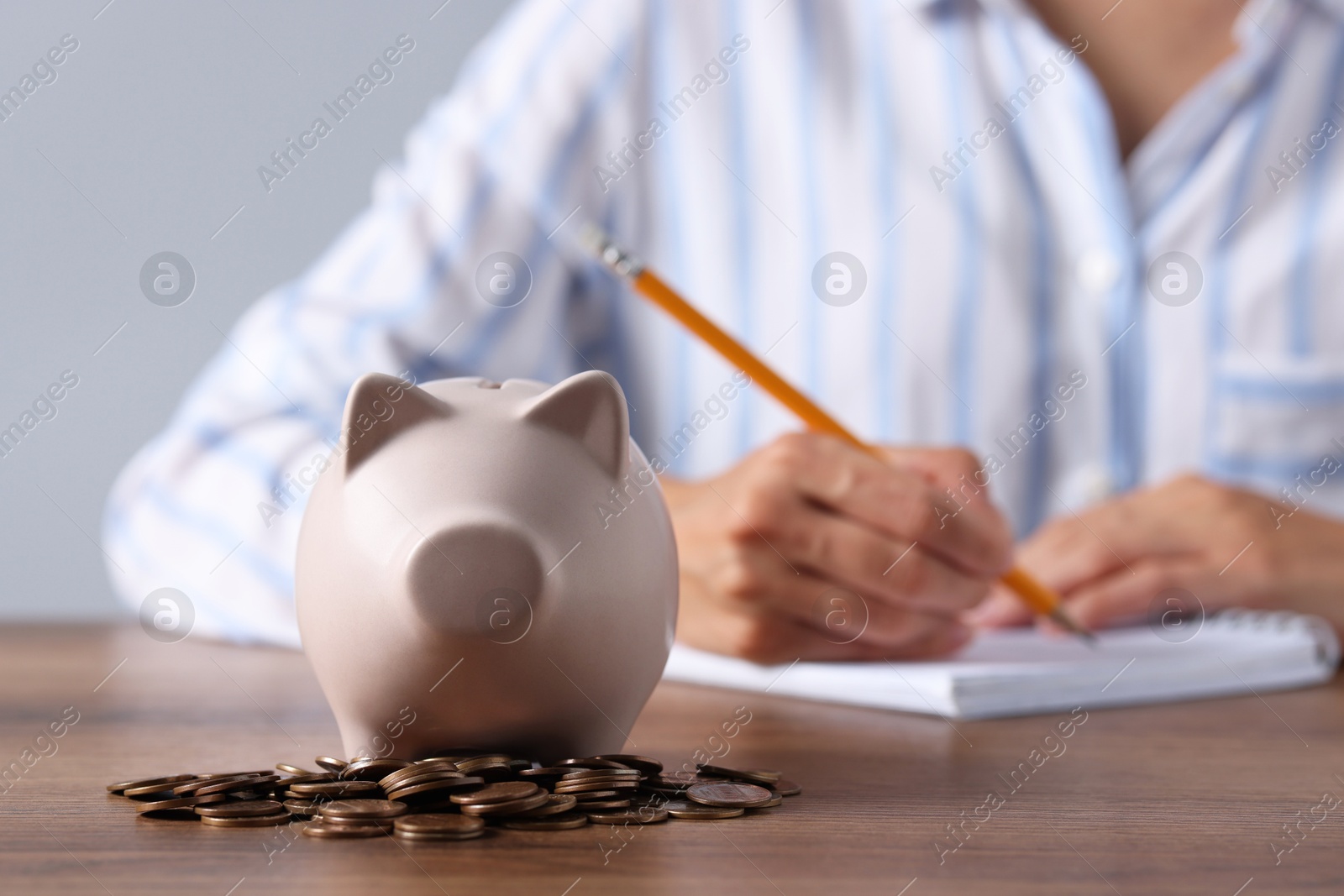 Photo of Woman taking notes at wooden table, focus on piggy bank and coins