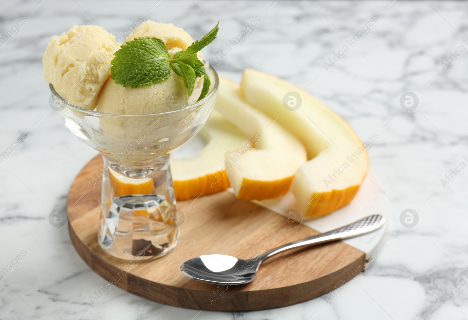 Photo of Scoops of melon sorbet with mint in glass dessert bowl, fresh fruit and spoon on light marble table, closeup