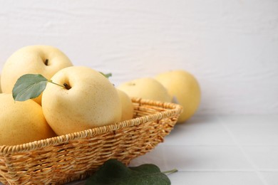 Photo of Delicious fresh apple pears in wicker basket and green leaves on white tiled table, closeup. Space for text