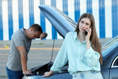 Photo of Man examining broken auto while woman calling to car service outdoors