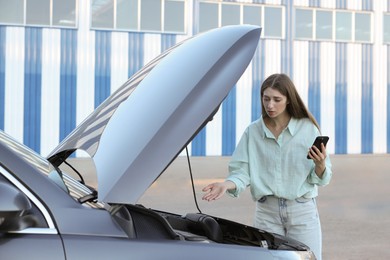 Stressed woman using smartphone near broken car outdoors