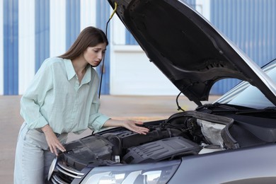 Stressed woman looking under hood of broken car outdoors