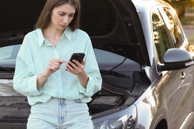 Stressed woman using smartphone near broken car outdoors