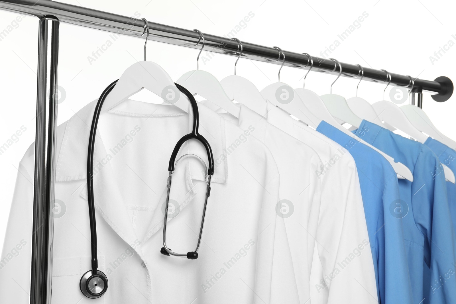 Photo of Different medical workers' uniforms and stethoscope on clothing rack against white background, closeup