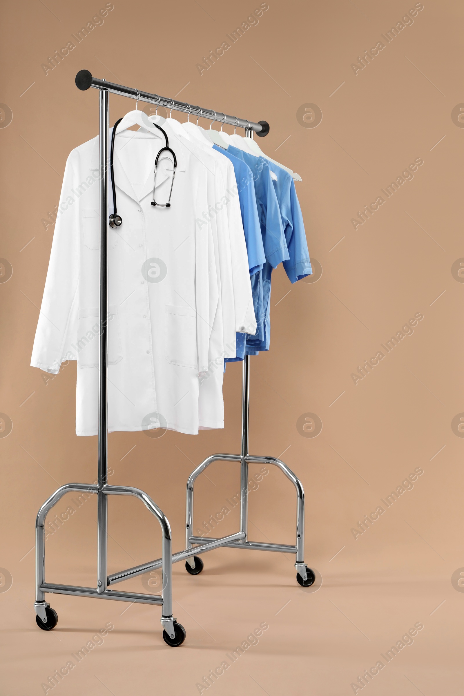 Photo of Different medical workers' uniforms and stethoscope on clothing rack against beige background