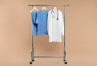 Photo of Different medical workers' uniforms and stethoscope on clothing rack against beige background