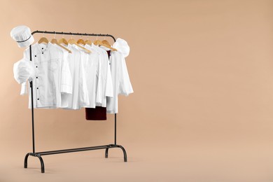 Photo of Different chef's uniforms on clothing rack against beige background, space for text