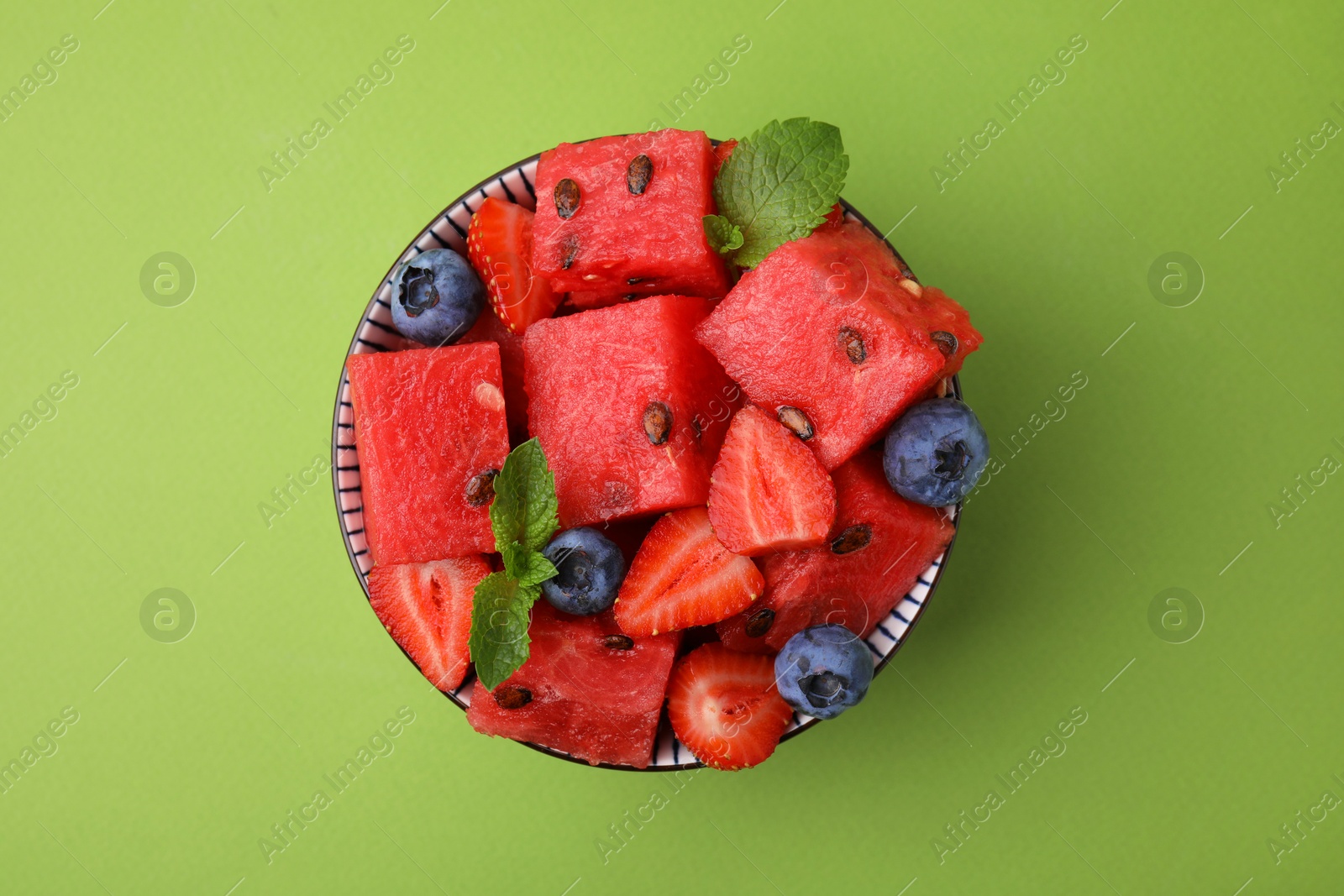 Photo of Pieces of tasty watermelon, strawberries, blueberries and mint in bowl on green table, top view