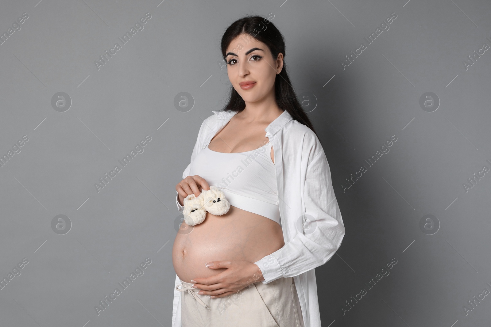 Photo of Portrait of beautiful pregnant woman with baby shoes on grey background