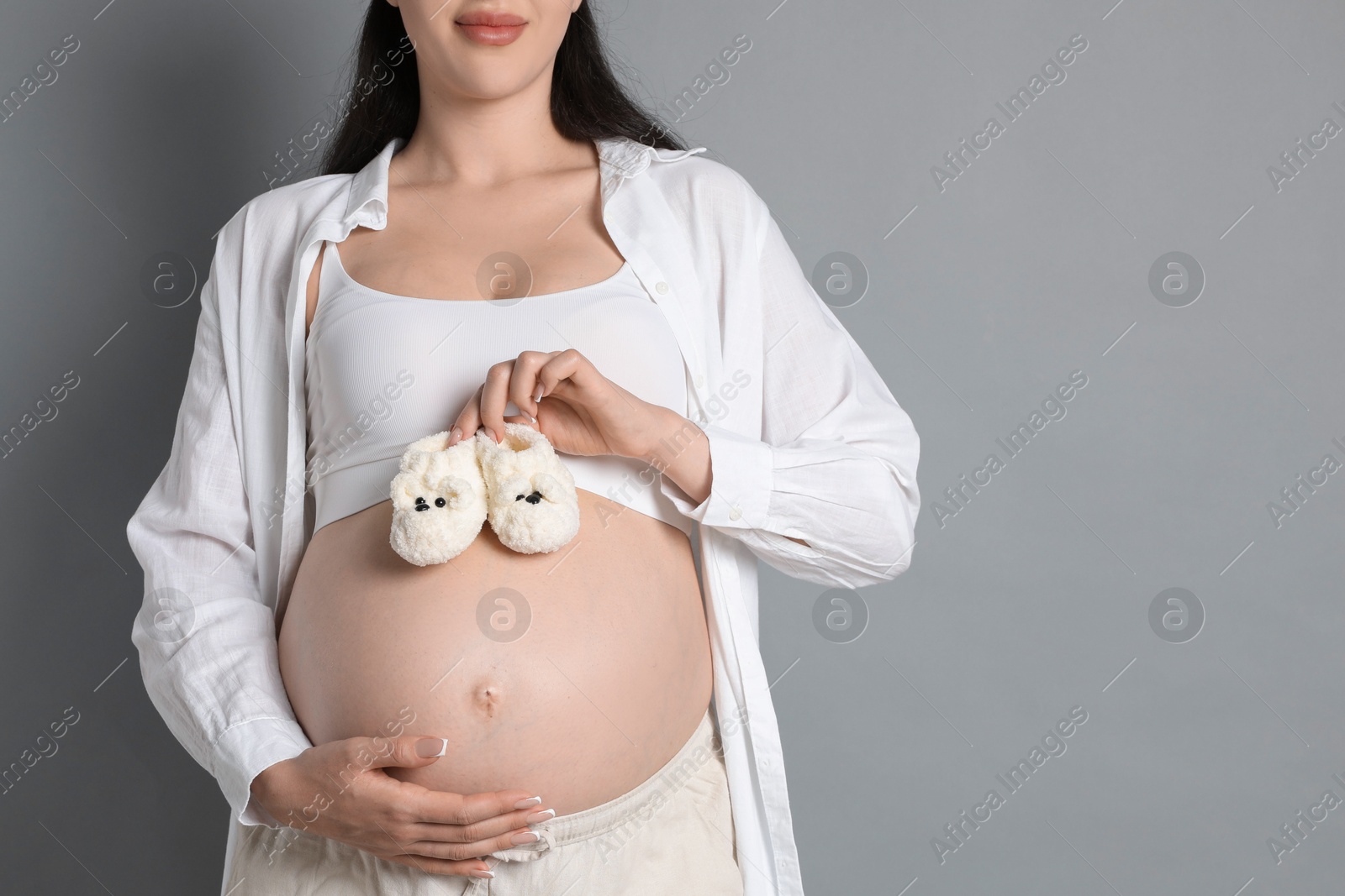 Photo of Young pregnant woman with pair of baby shoes on grey background, closeup. Space for text