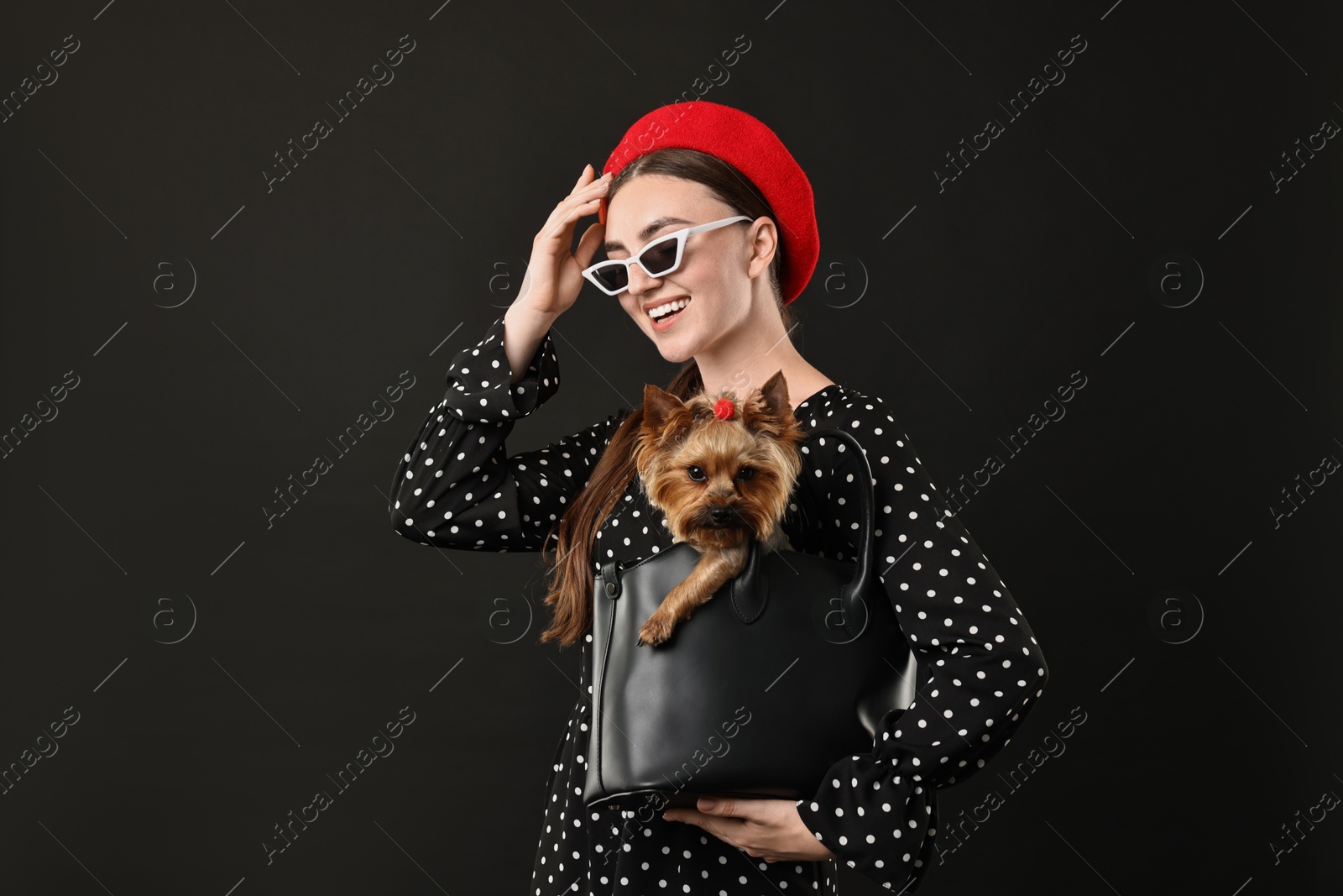 Photo of Woman in sunglasses holding bag with cute Yorkshire Terrier dog on black background