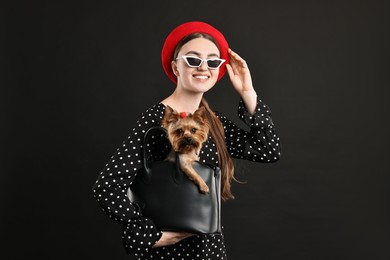 Woman in sunglasses holding bag with cute Yorkshire Terrier dog on black background