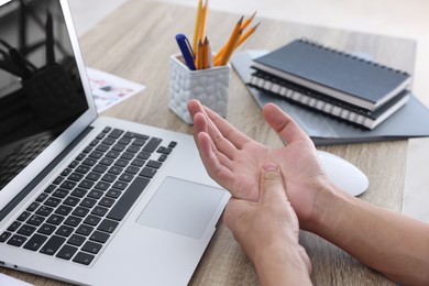 Photo of Man suffering from pain in wrist while working on laptop at table indoors, closeup. Carpal tunnel syndrome