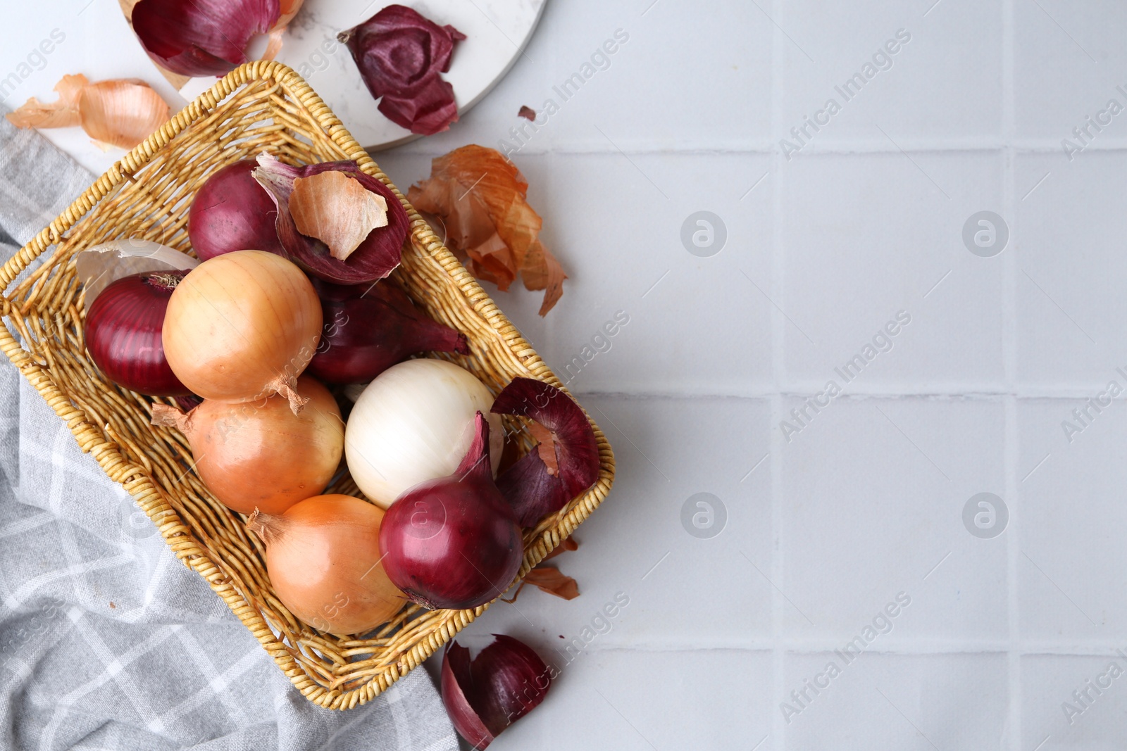 Photo of Wicker basket with fresh onions and peels on white tiled table, flat lay. Space for text