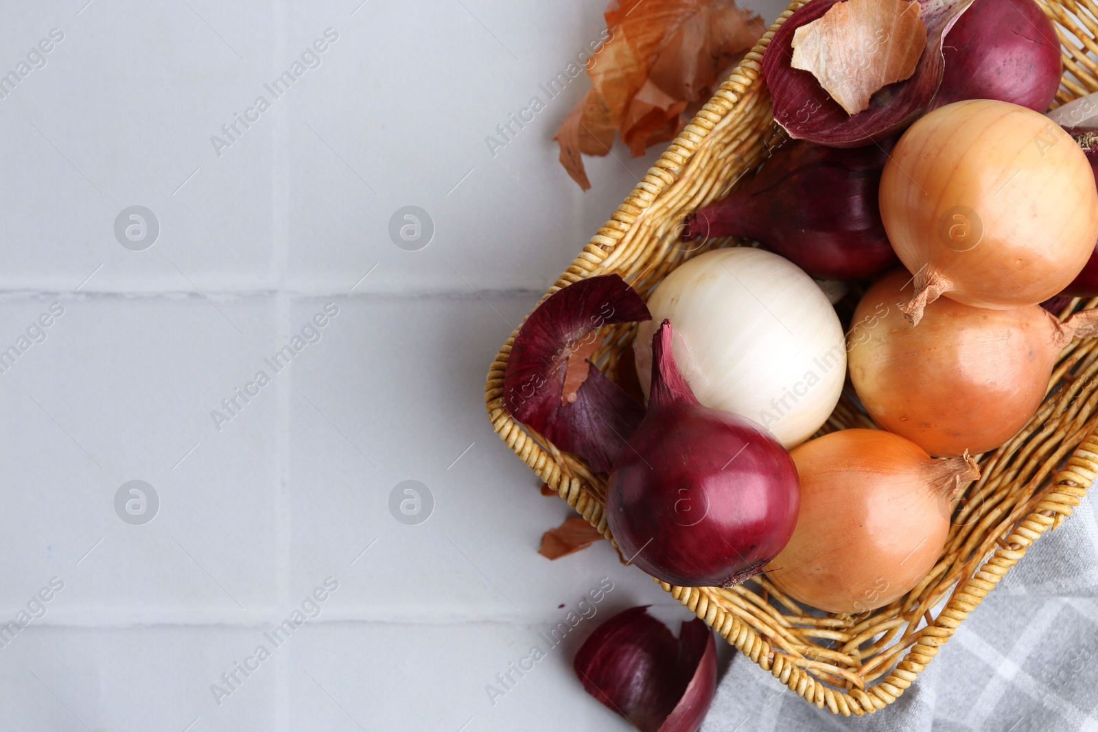 Photo of Wicker basket with fresh onions and peels on white tiled table, flat lay. Space for text