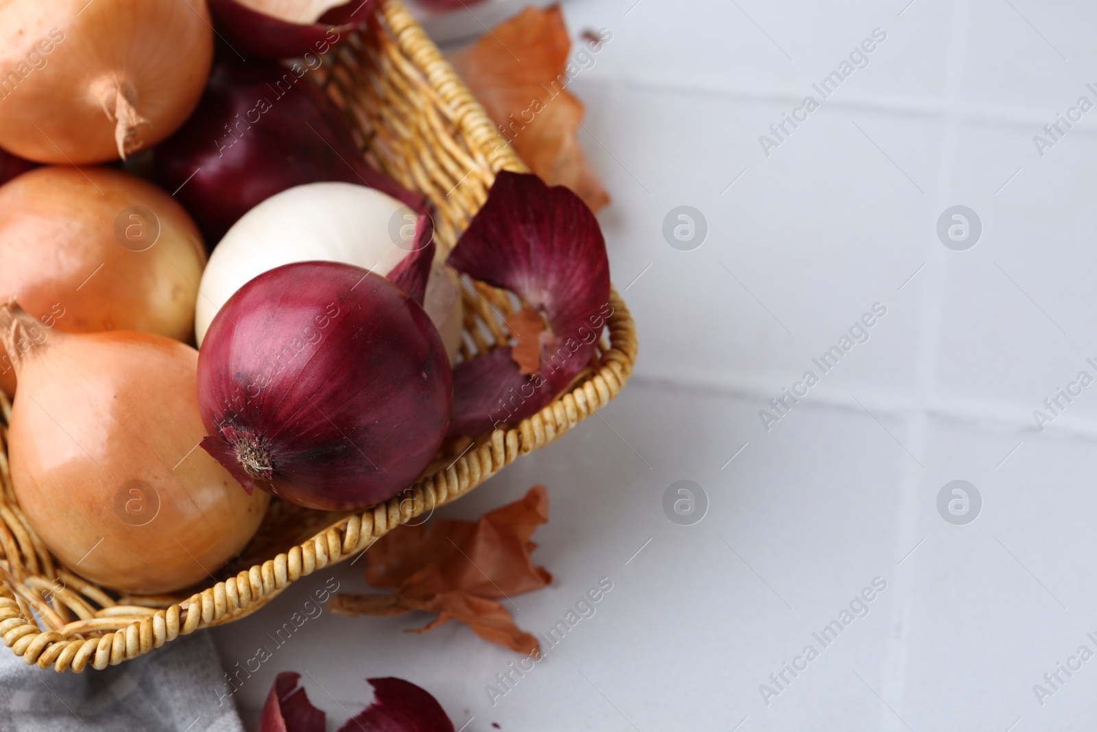 Photo of Wicker basket with fresh onions and peels on white tiled table, closeup. Space for text