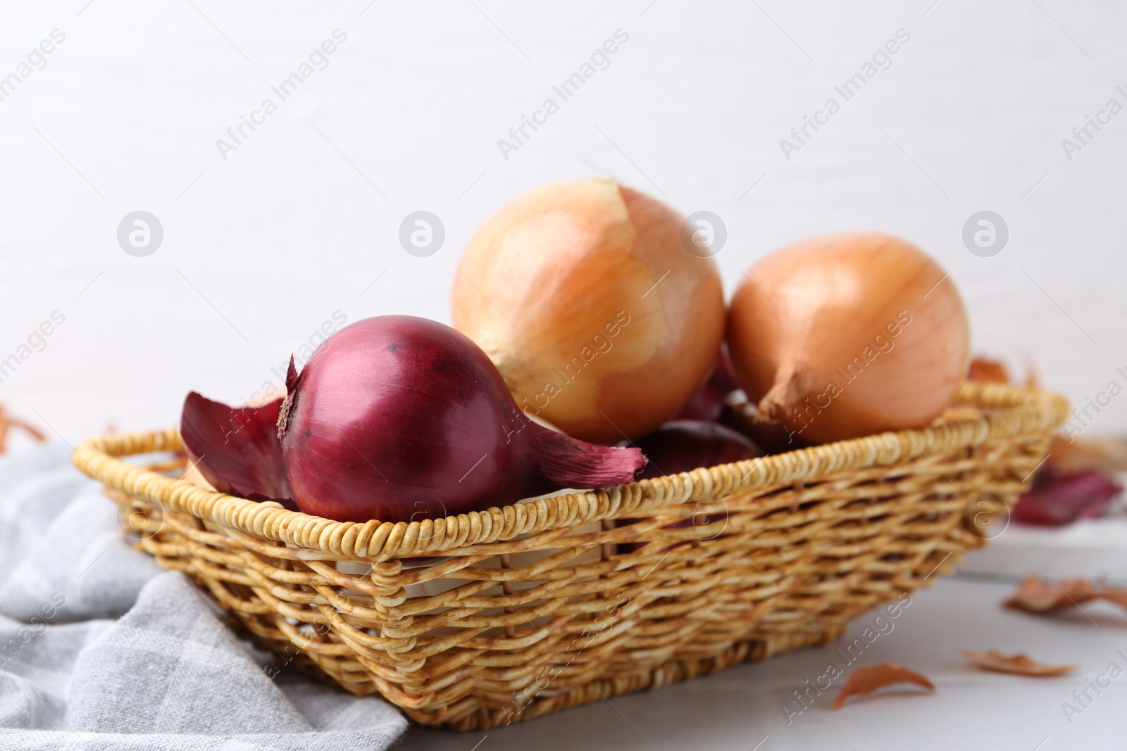 Photo of Wicker basket with fresh onions and peels on white tiled table, closeup