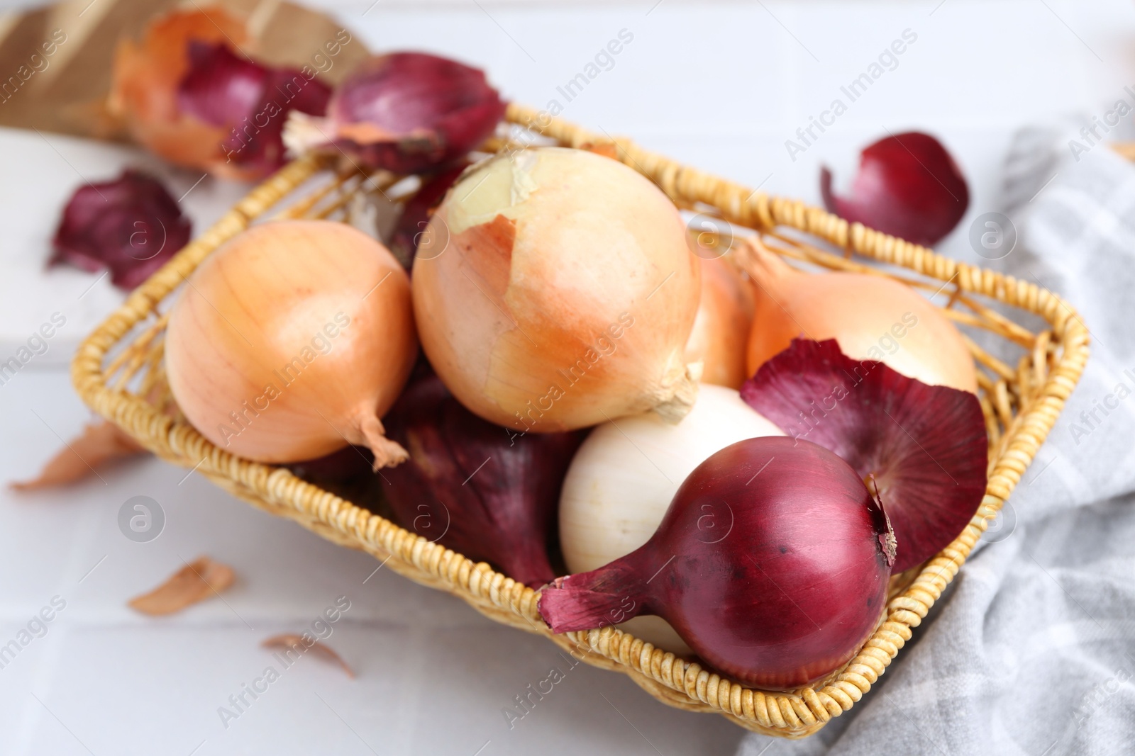 Photo of Wicker basket with fresh onions and peels on white tiled table, closeup