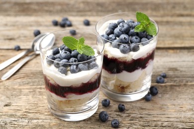 Photo of Tasty trifle dessert. Sponge cake, blueberries, jam and whipped cream in glasses on wooden table, closeup