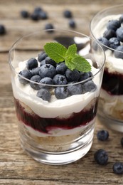 Photo of Tasty trifle dessert. Sponge cake, blueberries, jam and whipped cream in glasses on wooden table, closeup