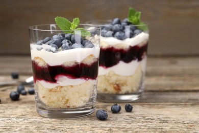 Photo of Tasty trifle dessert. Sponge cake, blueberries, jam and whipped cream in glasses on wooden table, closeup