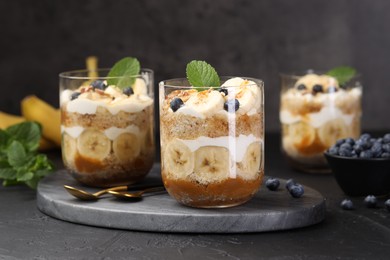 Photo of Tasty trifle dessert. Sponge cake, blueberries, banana and whipped cream in glasses on black table