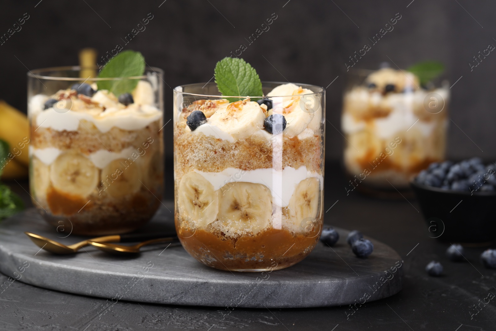 Photo of Tasty trifle dessert. Sponge cake, blueberries, banana and whipped cream in glasses on black table, closeup