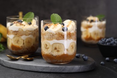 Photo of Tasty trifle dessert. Sponge cake, blueberries, banana and whipped cream in glasses on black table, closeup