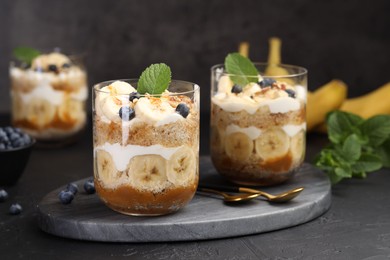 Photo of Tasty trifle dessert. Sponge cake, blueberries, banana and whipped cream in glasses on black table