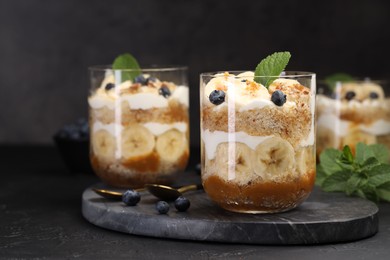 Photo of Tasty trifle dessert. Sponge cake, blueberries, banana and whipped cream in glasses on black table, closeup