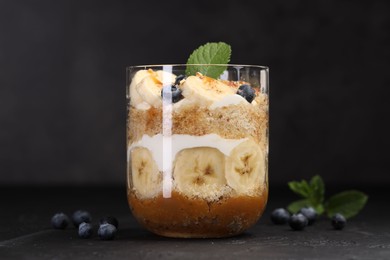 Photo of Tasty trifle dessert. Sponge cake, blueberries, banana and whipped cream in glass on black table, closeup